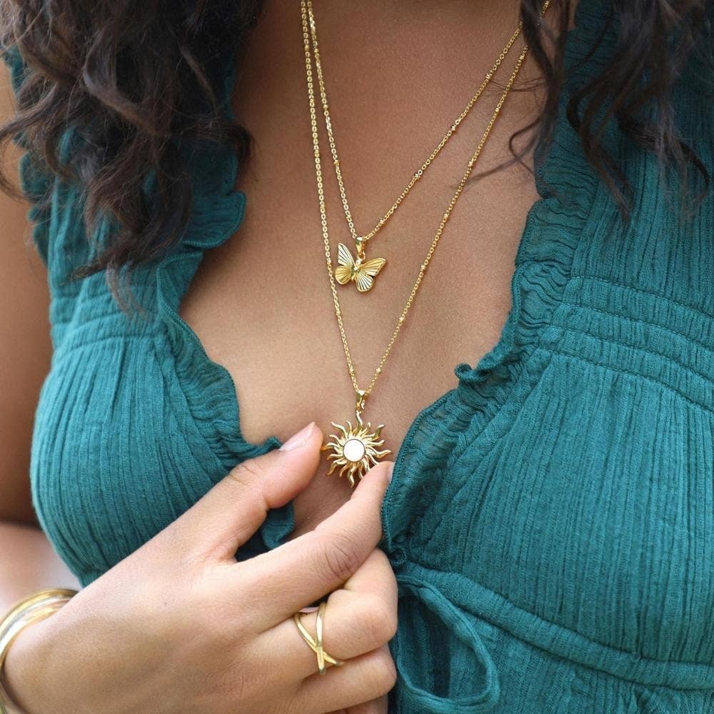 Woman wearing a teal dress with gold necklaces featuring butterfly and sun pendants.