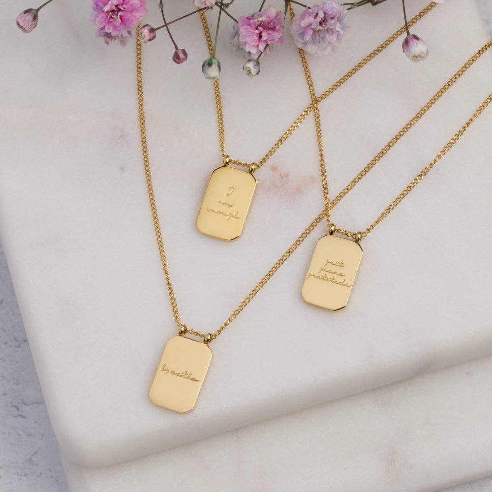 Three gold necklaces with engraved tags on a light background with pink flowers.