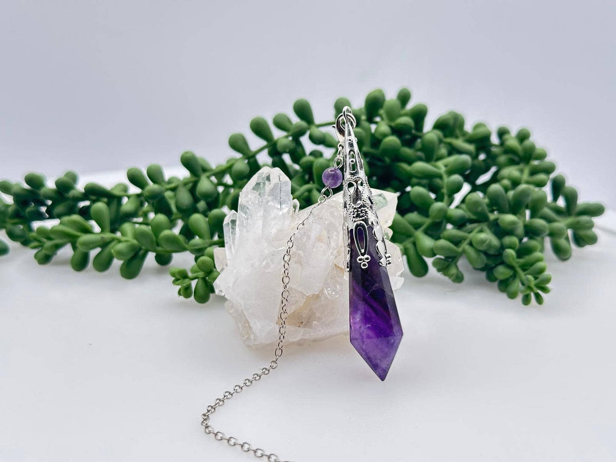 Purple crystal pendant on a chain with green foliage and a white crystal in the background