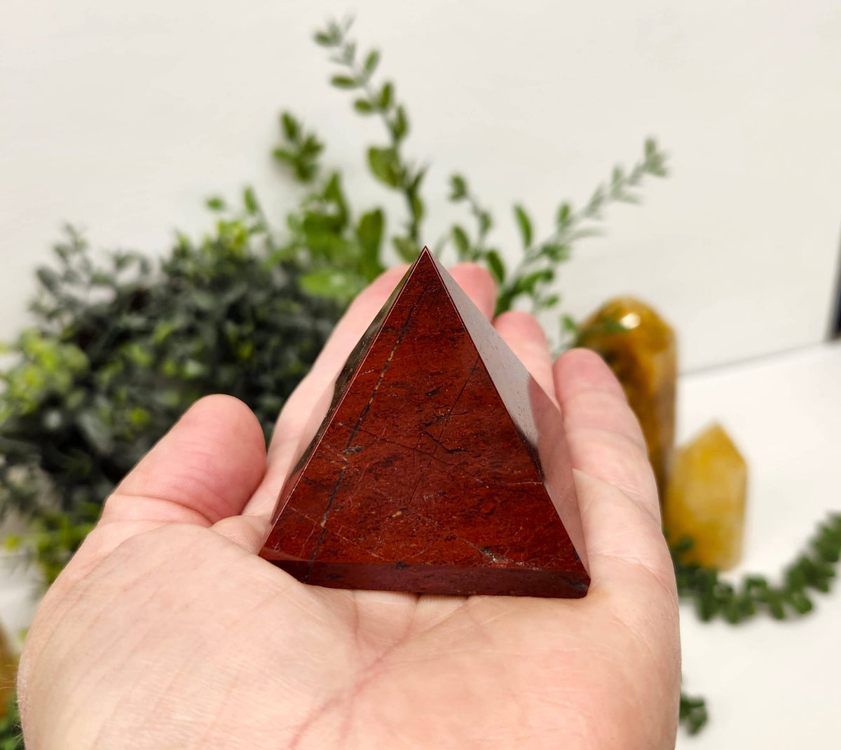 Hand holding a red pyramid crystal with a blurred background of greenery and another crystal.