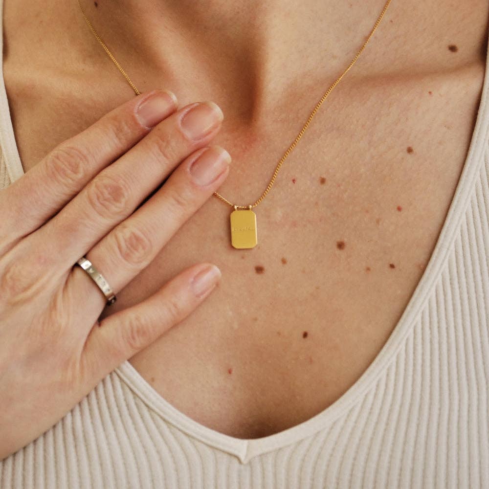 Gold necklace with a rectangular pendant worn by a person, with a hand touching the neck.