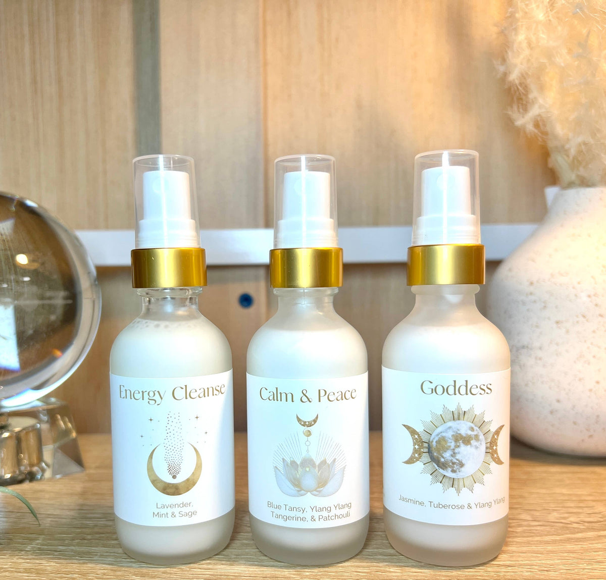 Three bottles labeled 'Energy Cleanse', 'Calm & Peace', and 'Goddess' on a wooden surface.
