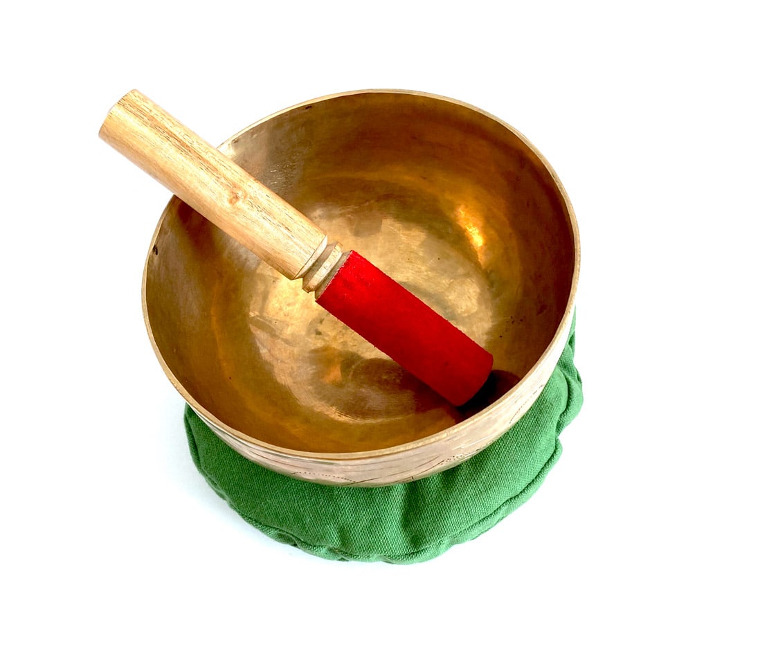 A gold-colored Himalayan singing bowl with a red striker, placed on a green cushion, on a white background - JOY - Journey Of You - Spiritual, Holistic, Empowering