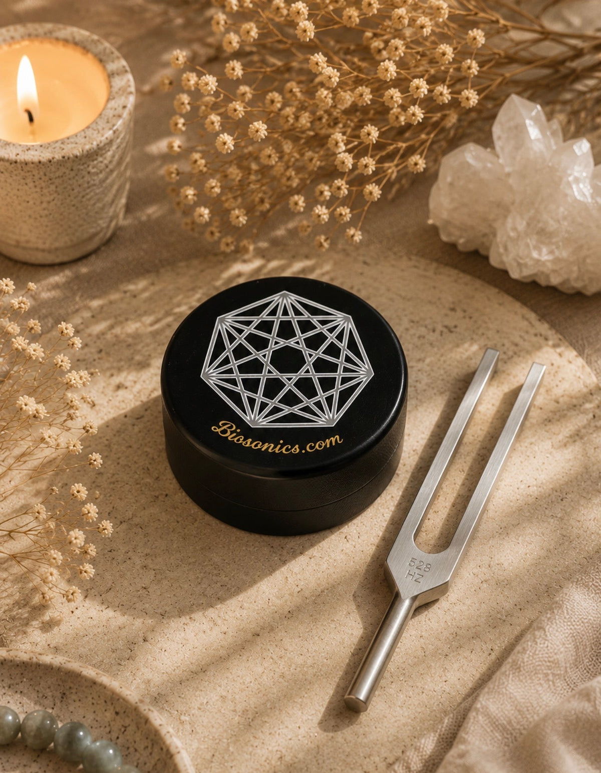 A black round Biosonics sound box container with a white geometric logo and gold website text, styled on a beige fabric surface with a silver tuning fork, dried flowers, a lit candle, and crystals.