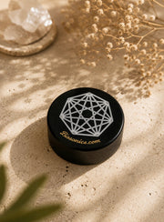 A round black jar lid with a white geometric line design and the text 'Biosonics.com' in gold, placed on a sandy surface with dried flowers and soft shadows.