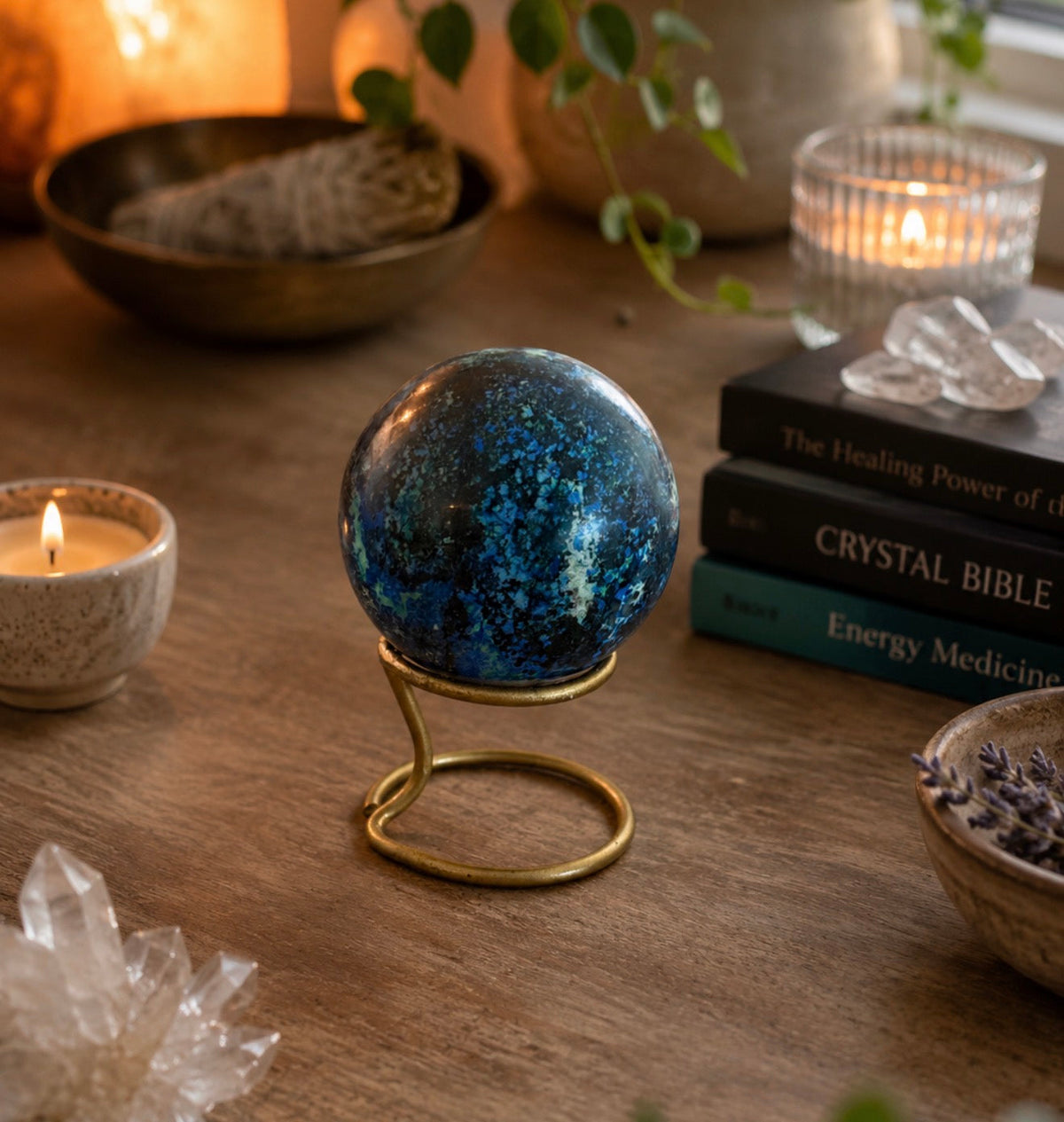 A polished blue crystal sphere on a small gold stand, displayed on a wooden table with candles, books, and crystals in a warm, styled setting.