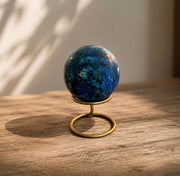 A polished blue lapis lazuli crystal sphere displayed on a small brass stand, set on a rustic wooden surface with warm natural light casting soft shadows.