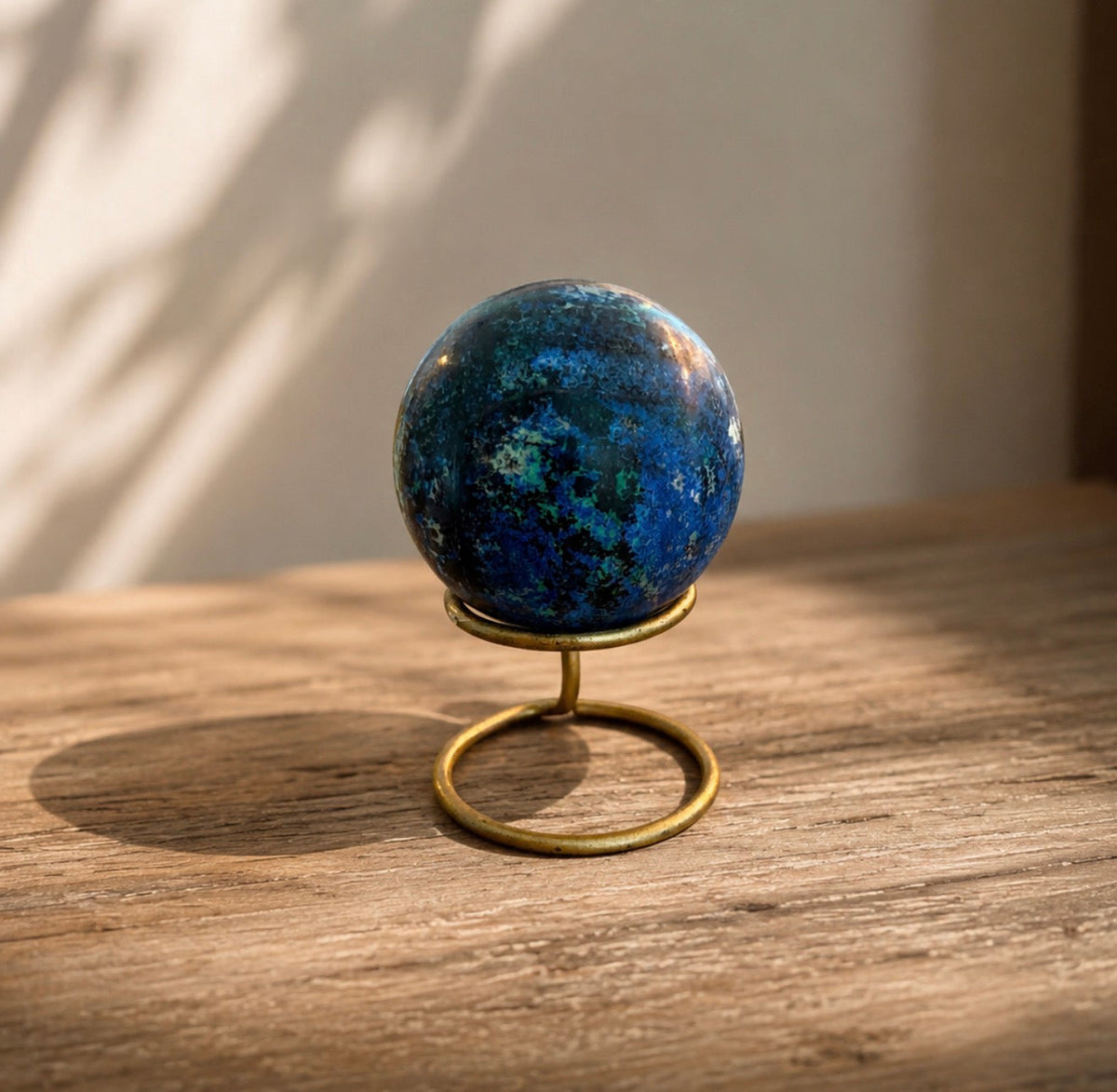 A polished blue lapis lazuli crystal sphere displayed on a small brass stand, set on a rustic wooden surface with warm natural light casting soft shadows.