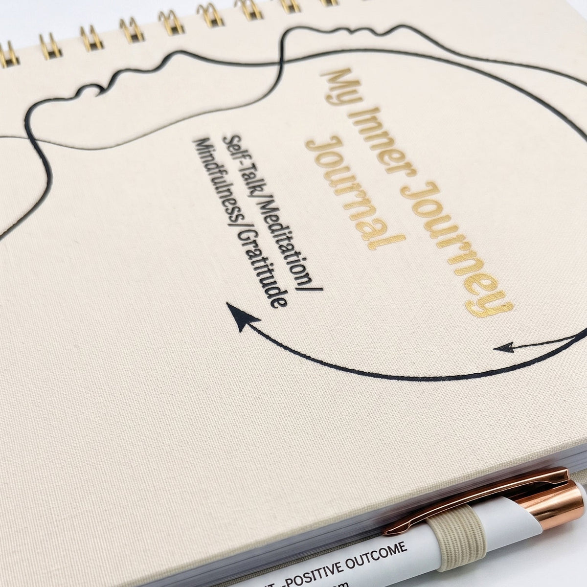 A close-up of a spiral-bound journal with a cream cover, featuring the title 'My Inner Journey Journal' in gold and black text, along with a pen resting across the bottom.