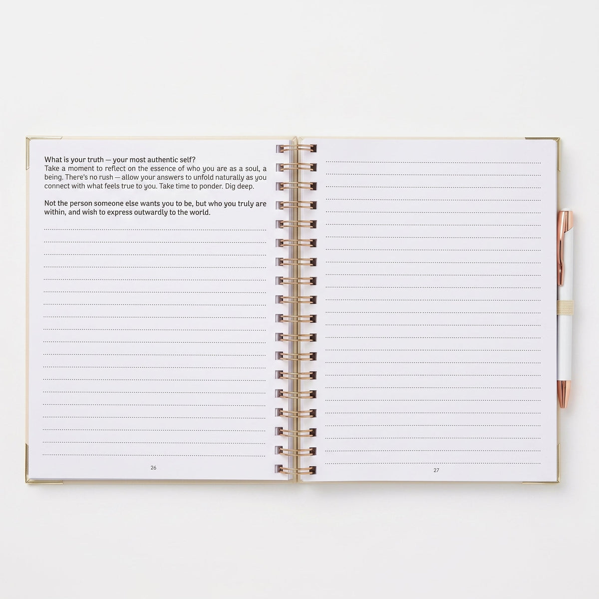 An open spiral-bound journal with lined pages, showing a reflective prompt on the left page and writing space on the right. A rose-gold pen is placed on the right side against a clean white background.