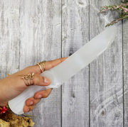 A hand holding a polished white selenite crystal knife against a wooden background with decorative elements - Moroccan Selenite Athame – Energy Knife for Cord Cutting &amp; Aura Clearing