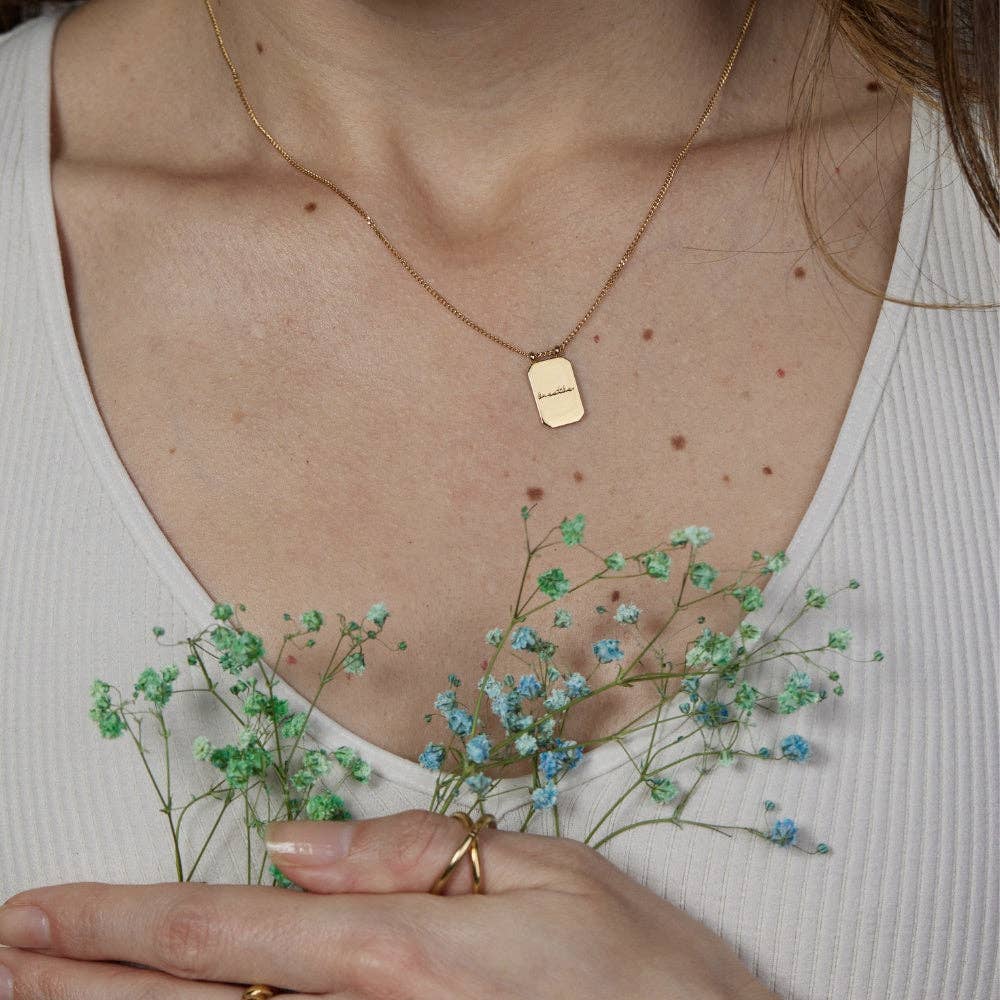 Person wearing a gold necklace with a tag, holding small green flowers.