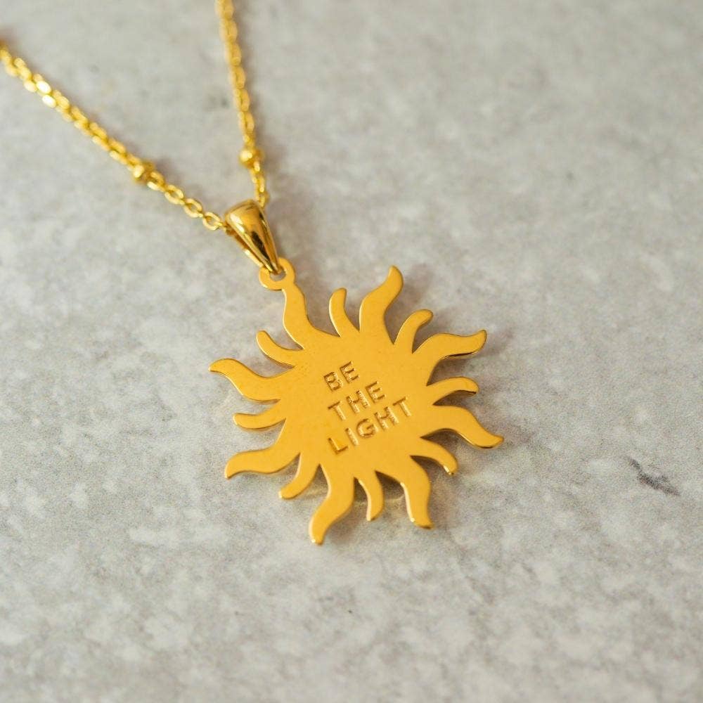 Gold sun-shaped pendant necklace with 'Be The Light' engraving on a light gray background
